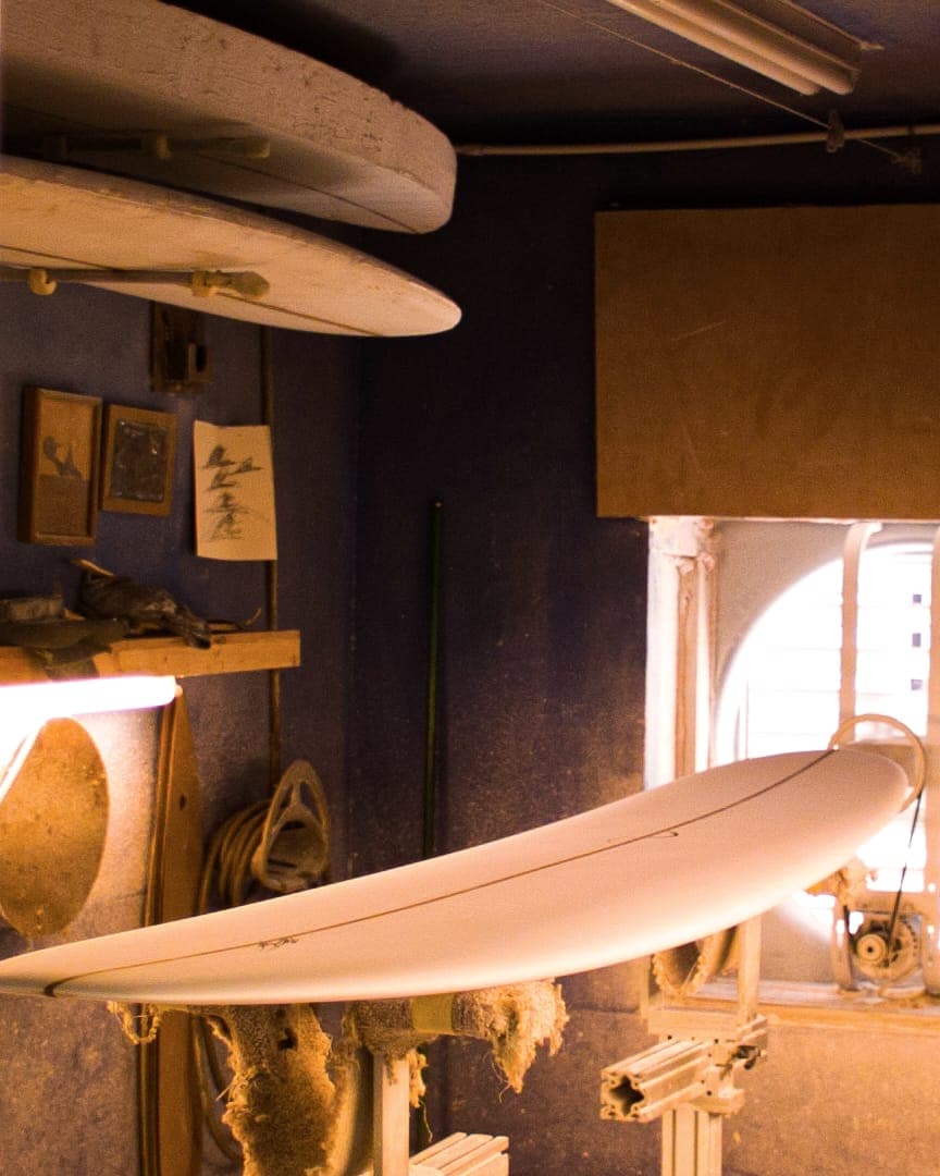 A surfboard on stands in a shaping bay, lit through a round window.
